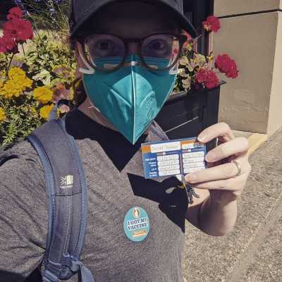 Me, wearing an N95 and black baseball hat, holding up my vaccine record.